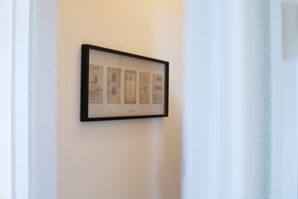Im Vordergrund ein Türrahmen, dahinter an einer Wand, ein schwarzer Rahmen mit fünf Bildern von Leonardo da Vinci.